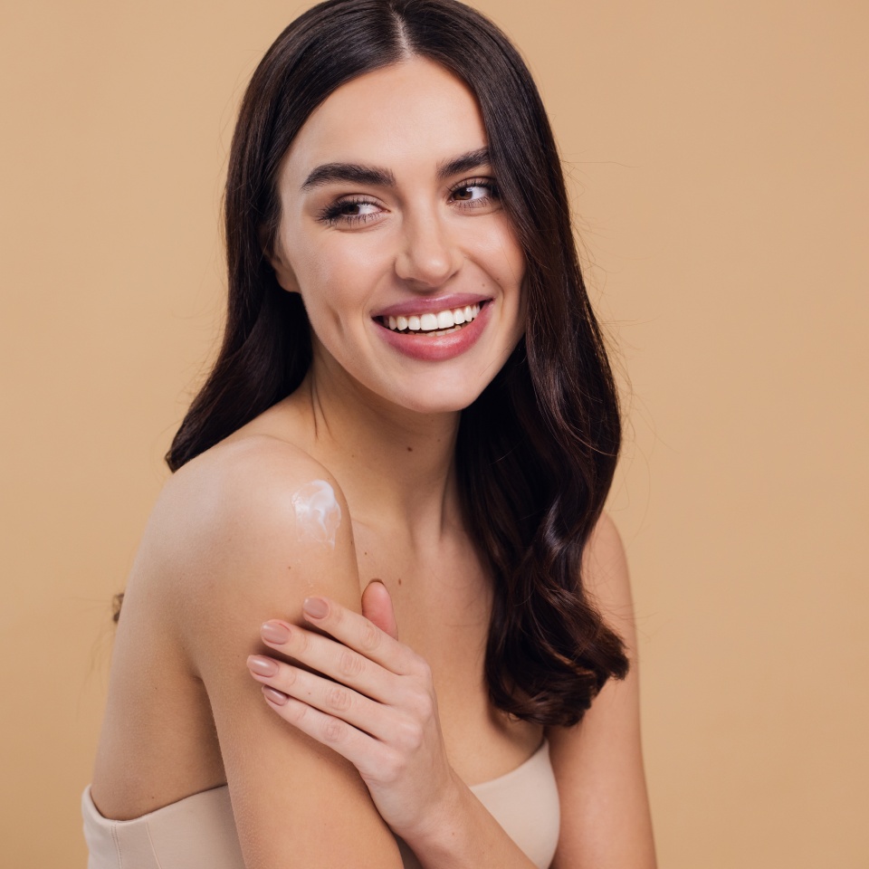 Close up beauty portrait of a laughing beautiful half naked woman smiling and applying face cream isolated over beige background