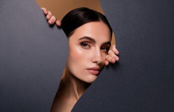 A woman with dark hair and neutral makeup looks through a jagged opening in a dark sheet of paper, holding the edges with both hands.